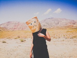 mujer en desierto con bolsa de papel en la cabeza que tiene dibujada una mueca triste